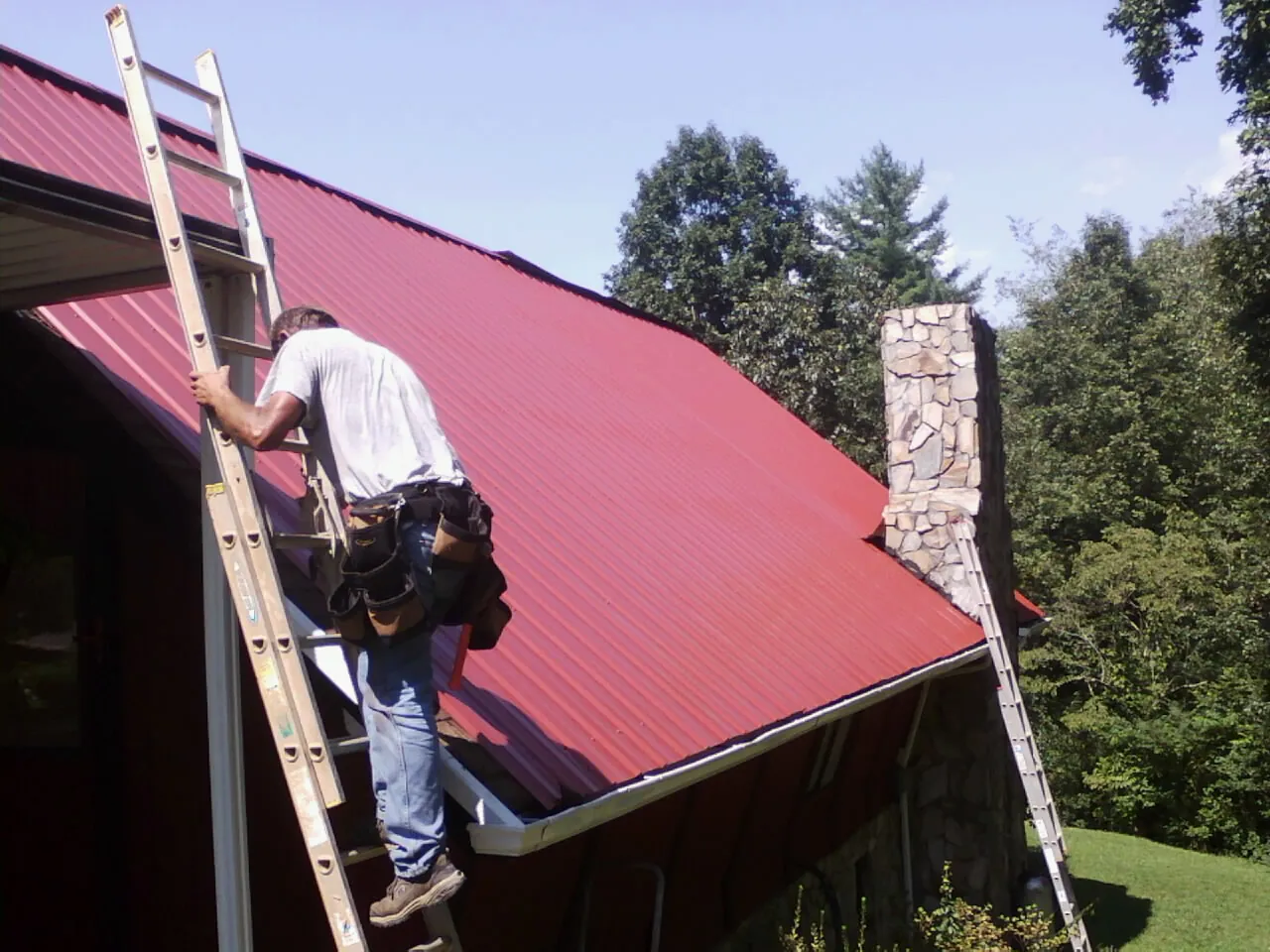 Roofer on a ladder beside a red metal roof and stone chimney — Soffit & Fascia Repair by Roofing Brothers in Hacker Valley